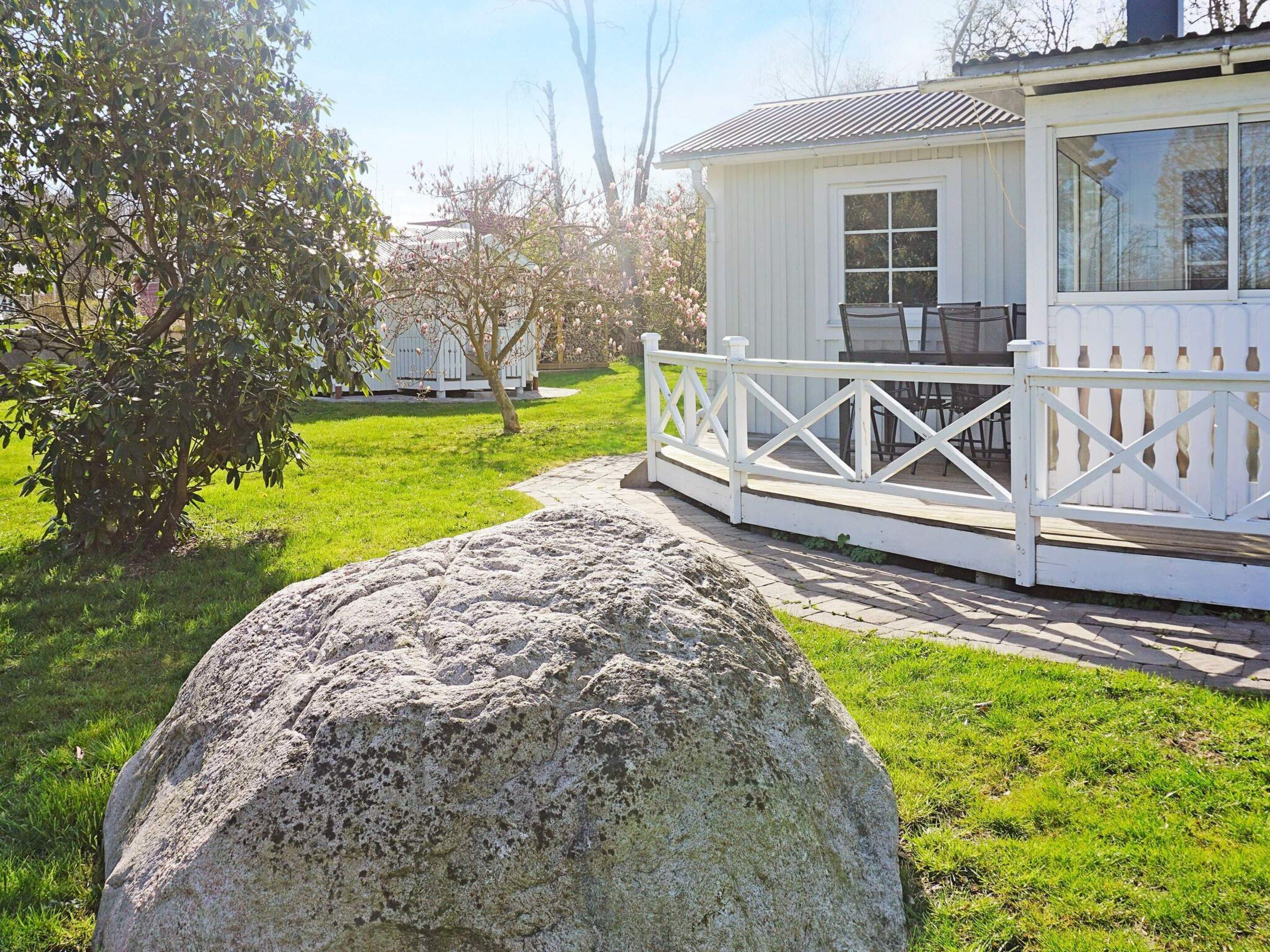 A white wooden house with a porch, a large rock in the foreground, and a tree with pink blossoms.