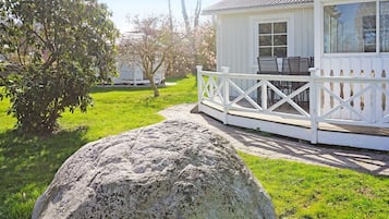 A white wooden house with a porch, a large rock in the foreground, and a tree with pink blossoms.