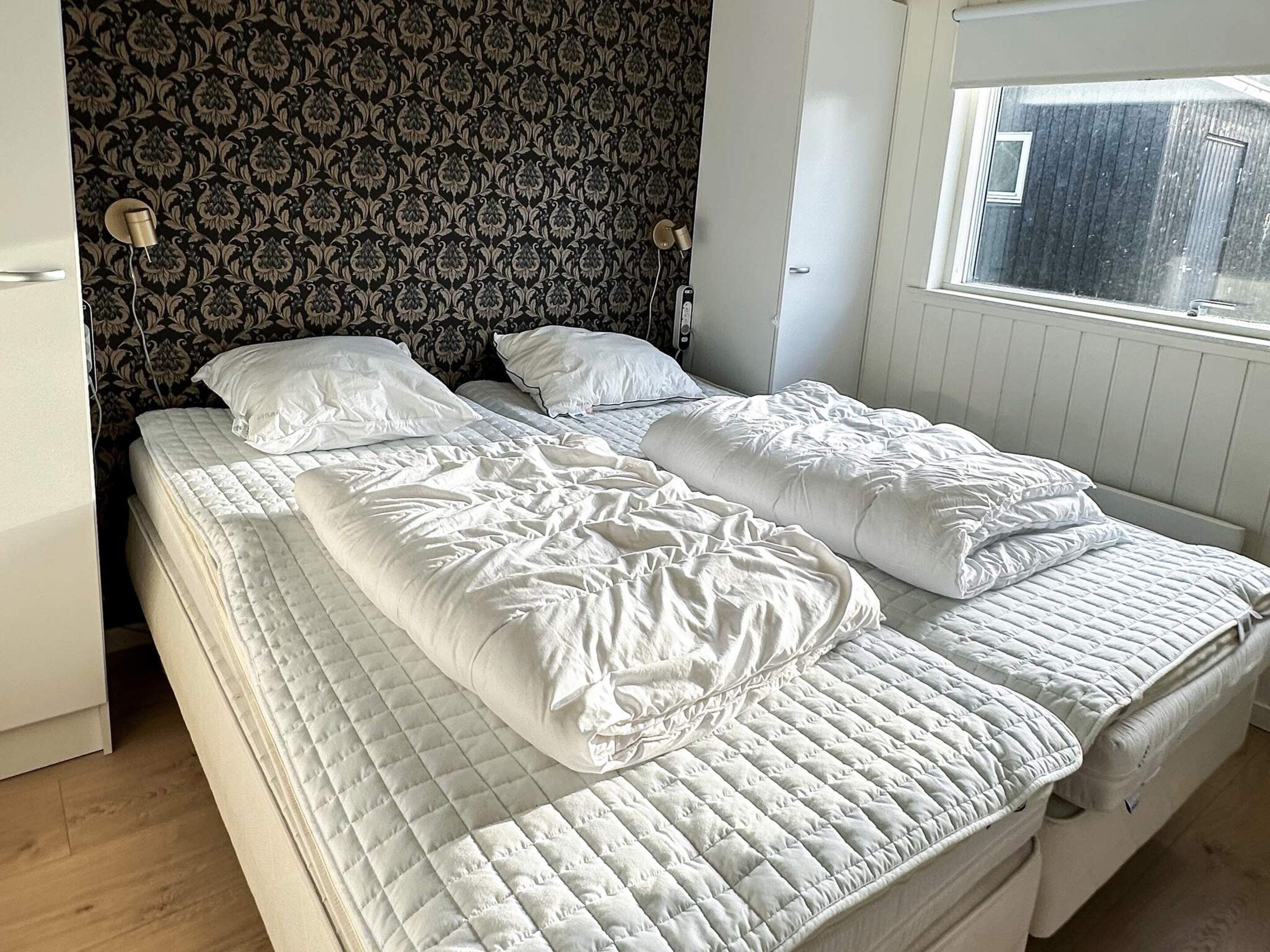 A bedroom with two beds, a patterned wallpaper, a window, and a white door.