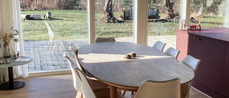 Un comedor moderno con una gran mesa redonda, sillas blancas y vistas a un jardín y árboles a través de amplios ventanales.