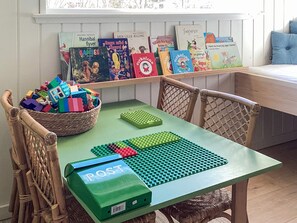 Un espace de jeux pour enfants avec une table, des chaises, un panier rempli de blocs de construction colorés et une collection de livres pour enfants sur une étagère.
