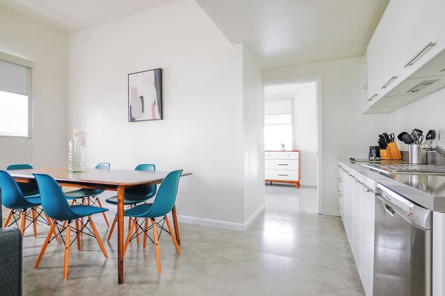 A modern kitchen with white cabinets, a stainless steel sink, and a dining area with a wooden table and blue chairs.