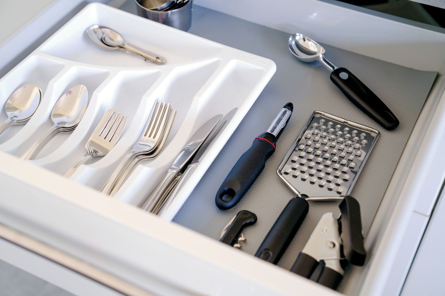 A drawer containing various kitchen utensils including spoons, forks, knives, a grater, and a peeler.