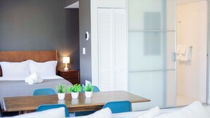 Moderni hotellihuone, jossa on sänky, yöpöytä, lamppu, ruukkukasveja sekä ruokailutila, jossa on tuoleja ja pöytä.