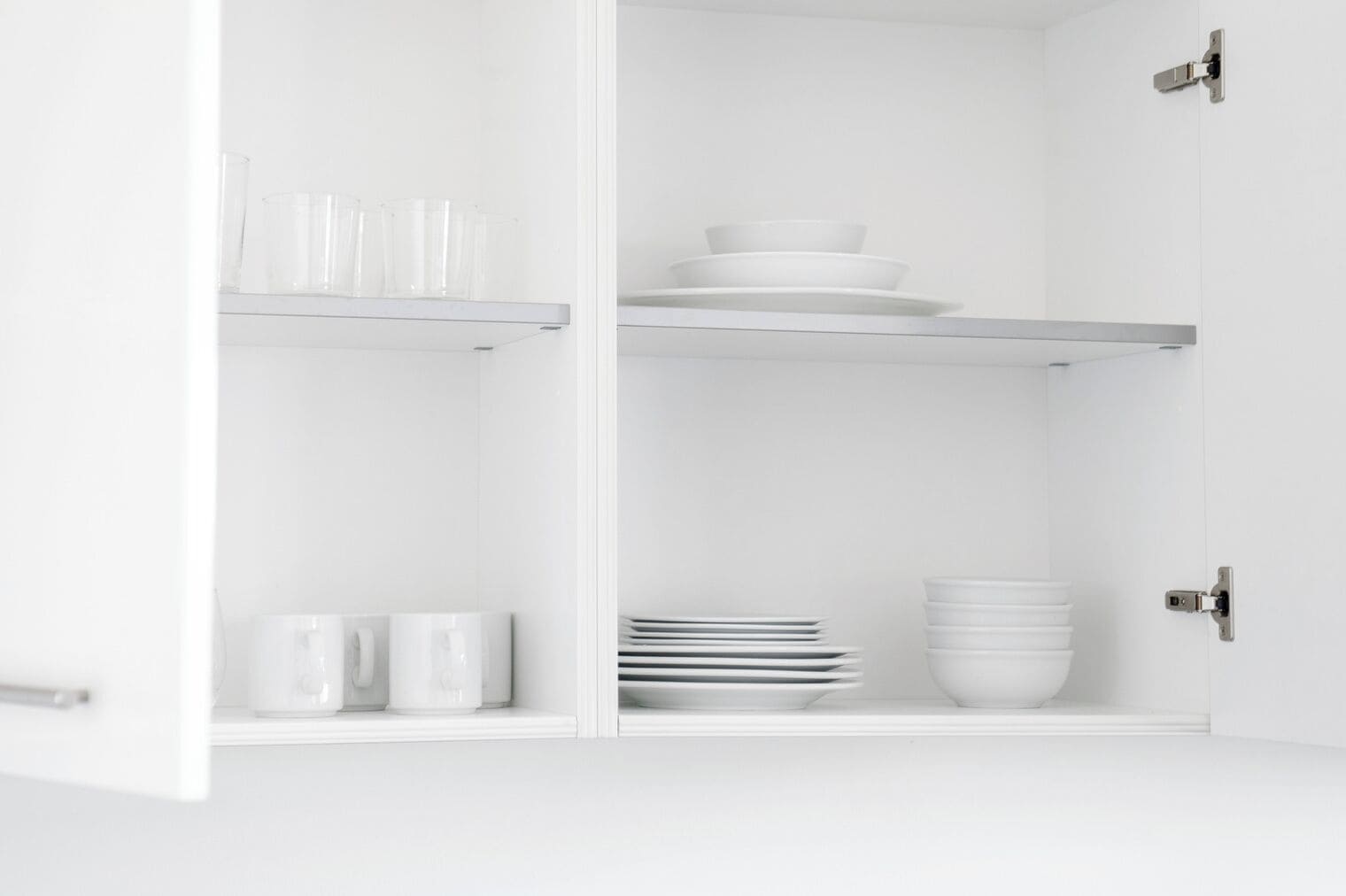 A white cabinet with shelves holding clear glasses, white plates, and white bowls.