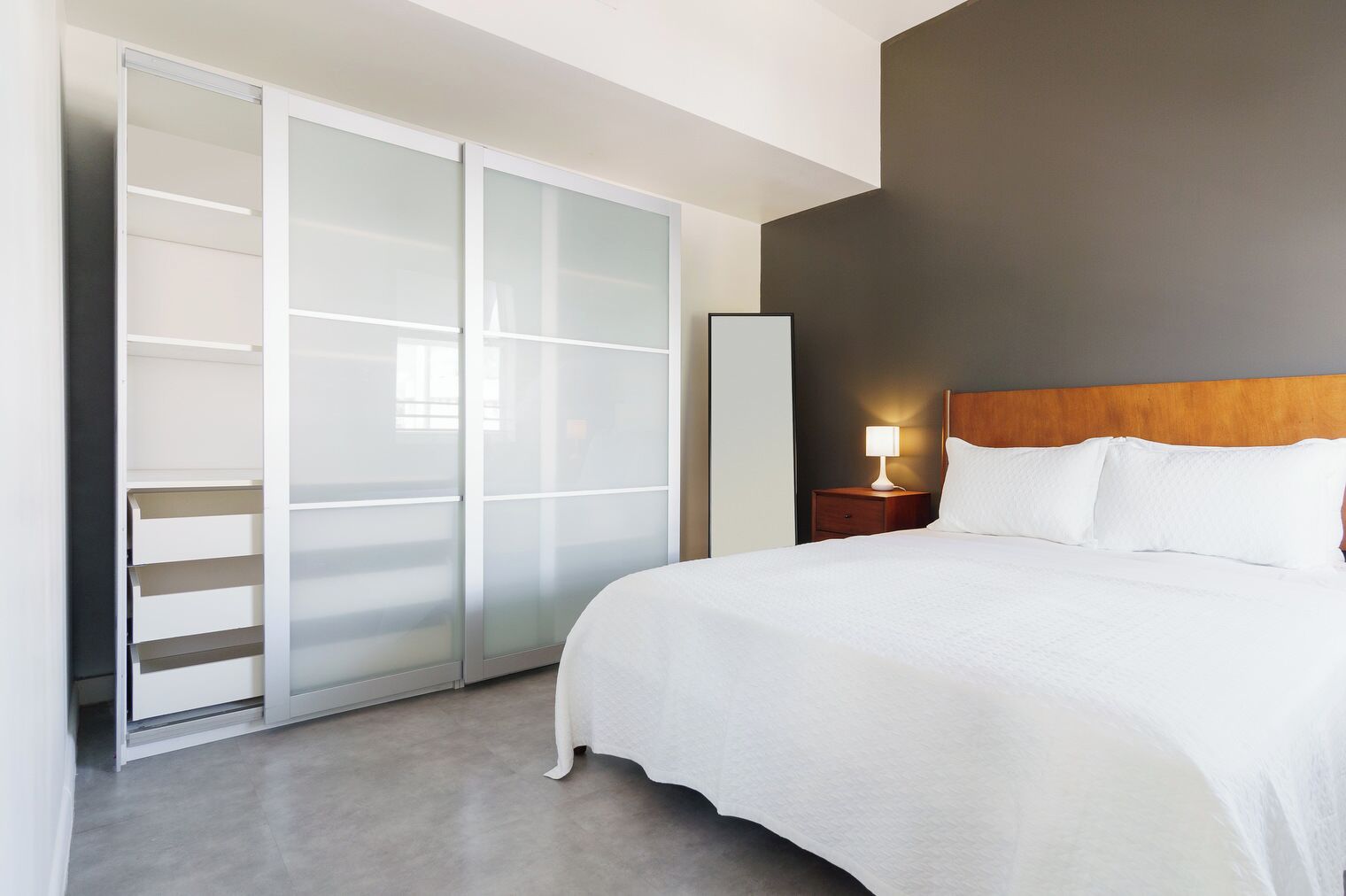 A hotel room with a bed, a nightstand with a lamp, a wardrobe with sliding doors, and a dark accent wall.