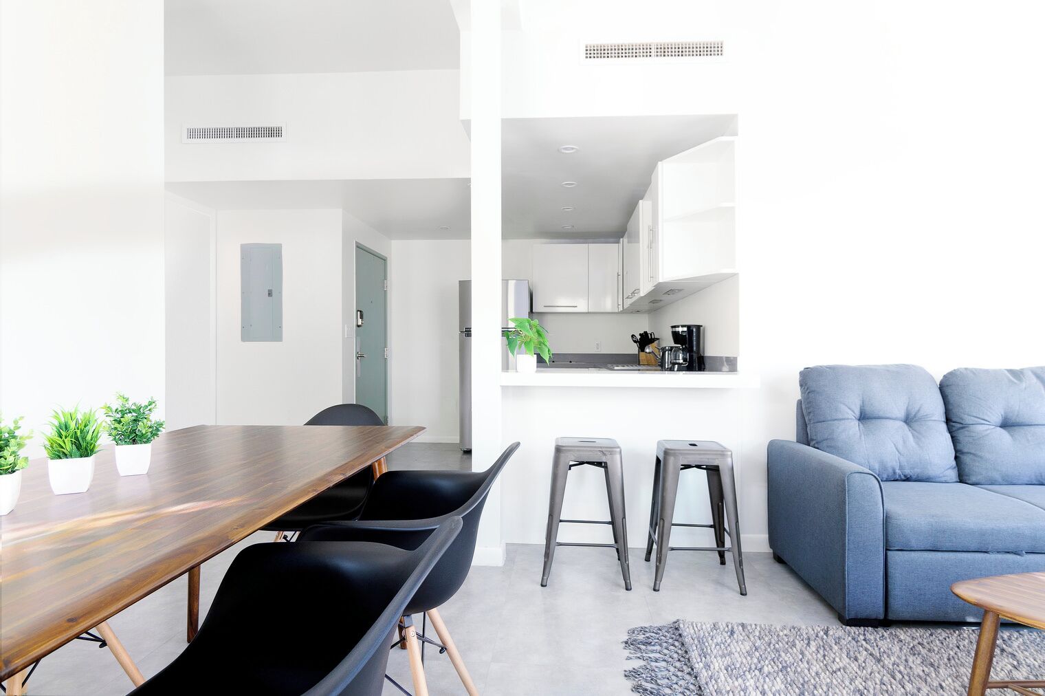 Un espacio habitable moderno con una mesa de comedor de madera, sillas negras, un sofá azul y una zona de cocina con armarios blancos y una cafetera.