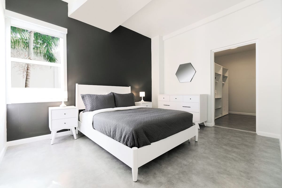 A modern bedroom with a large bed, white nightstands, a dresser, and a walk-in closet.
