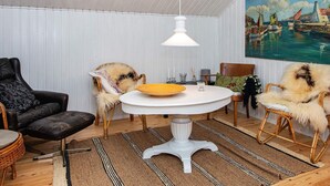 A room with a white ceiling, a round white table with a yellow plate, two wicker chairs with fur throws, a black leather sofa, and a painting of a harbor scene. - Tranquil Haven in Knebel (Knebel)