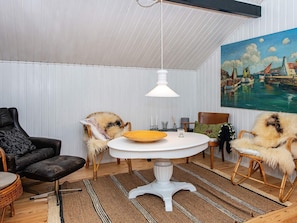A room with a white ceiling, a round white table with a yellow plate, two wicker chairs with fur throws, a black leather sofa, and a painting of a harbor scene. - 6 Person Holiday Home in Knebel (Knebel)