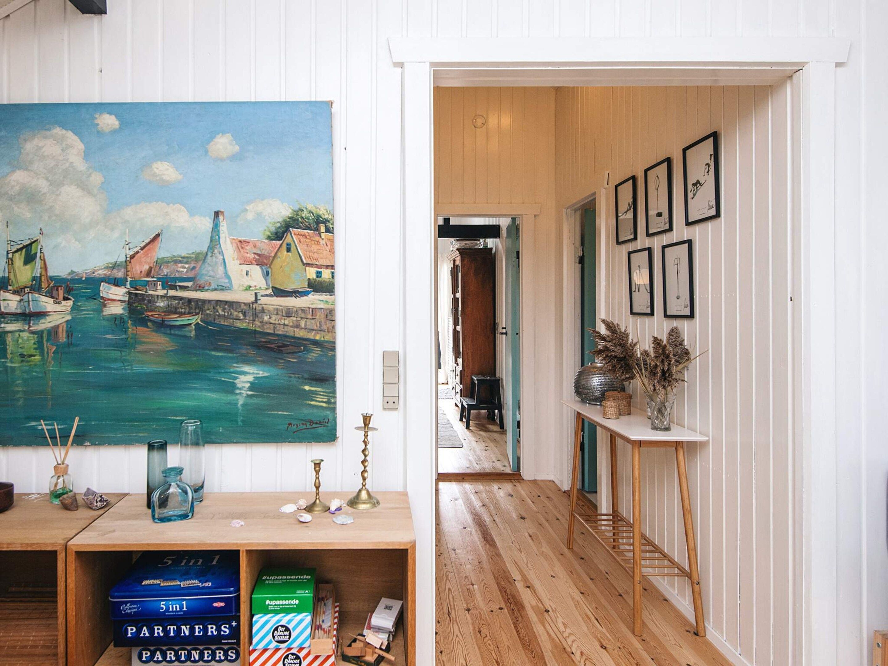 Un couloir doté d’un plancher en bois, d’un tableau représentant un paysage portuaire et d’une étagère sur laquelle se trouvent des boîtes et une lampe.