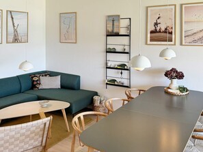 A modern living room with a green sofa, a round dining table, and a white coffee table. There are framed pictures on the wall, a black shelving unit with decorative items, and a white pendant light over the table.