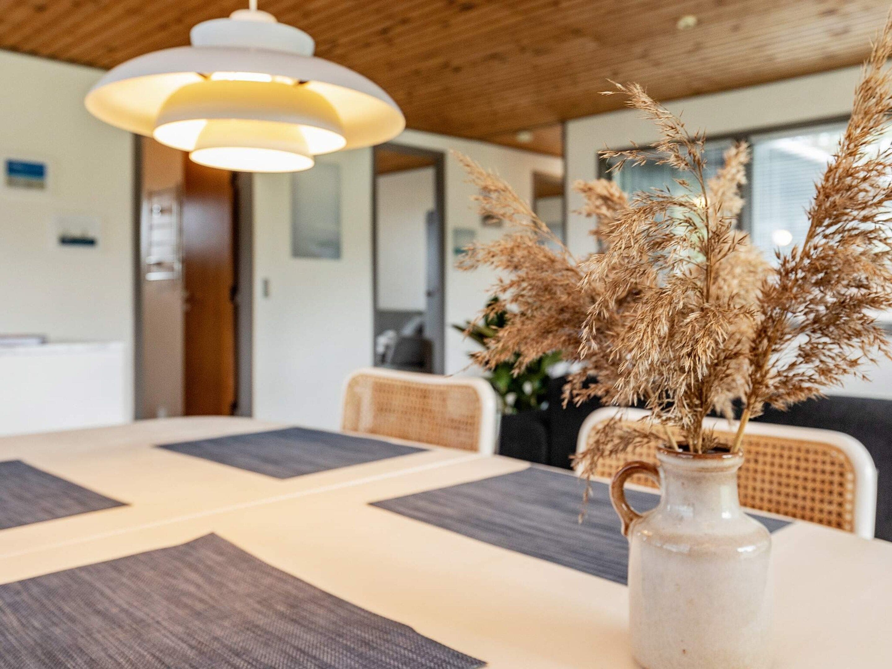 A dining area with a table, chairs, a vase with dried plants, and a hanging light fixture.