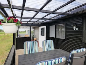 Une terrasse extérieure avec une table et des chaises, un pot de fleurs suspendu et une vue sur l’intérieur de la maison.