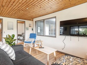 Un salon moderne avec un plafond en bois, un canapé gris, un fauteuil bleu, une petite table et une grande fenêtre avec des stores.