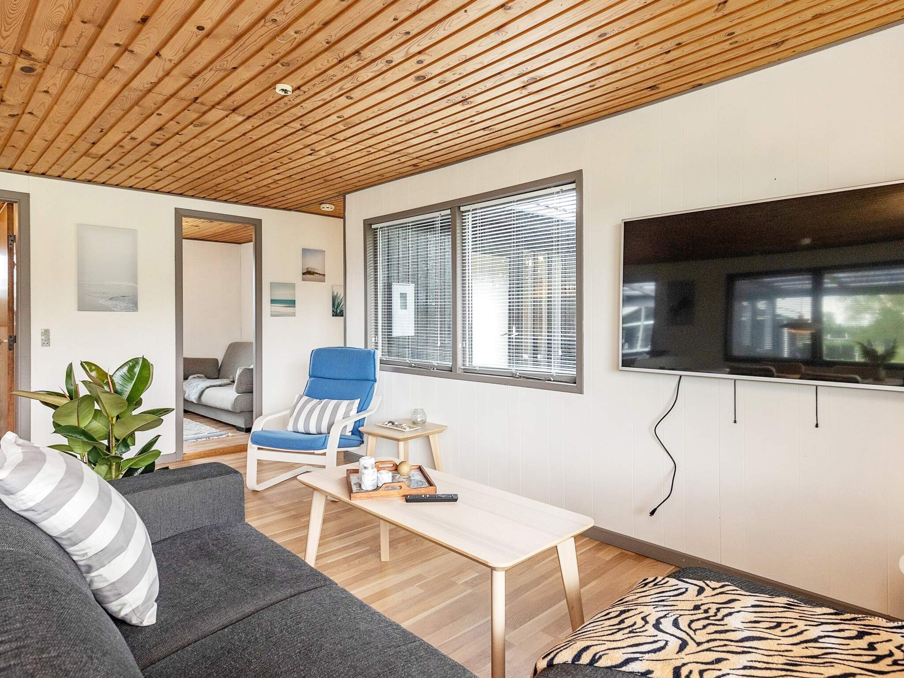 A modern living room with a wooden ceiling, a grey sofa, a blue armchair, a small wooden table, and a large window with blinds.