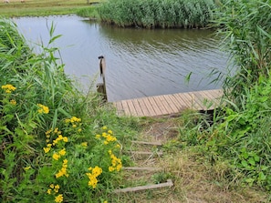 A wooden dock extending into a calm body of water, surrounded by lush greenery and yellow flowers. - 5 Person Holiday Home in Saeby (Sæby)
