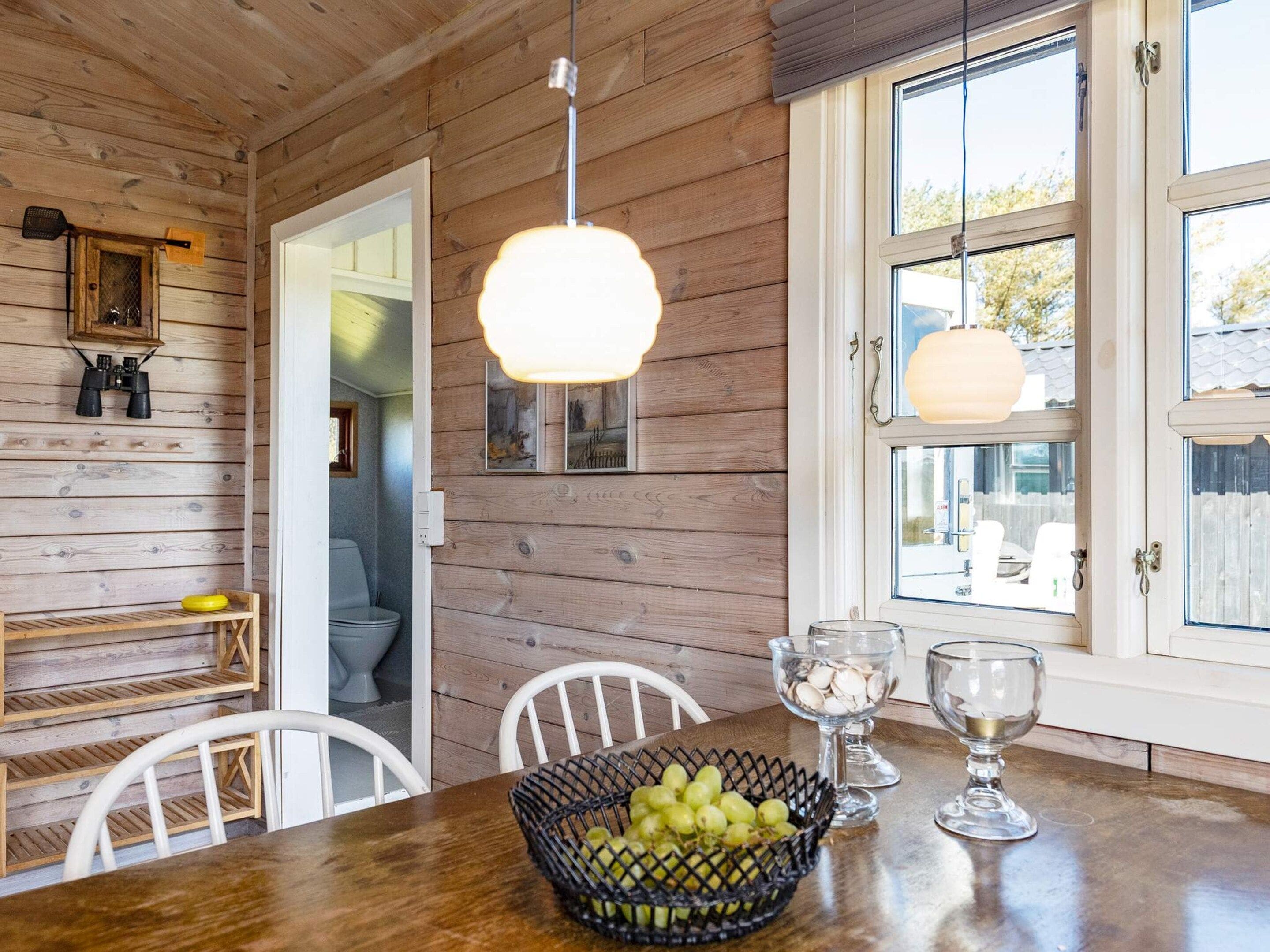 A wooden interior with a dining table, chairs, a basket of fruit, and a window with a view.