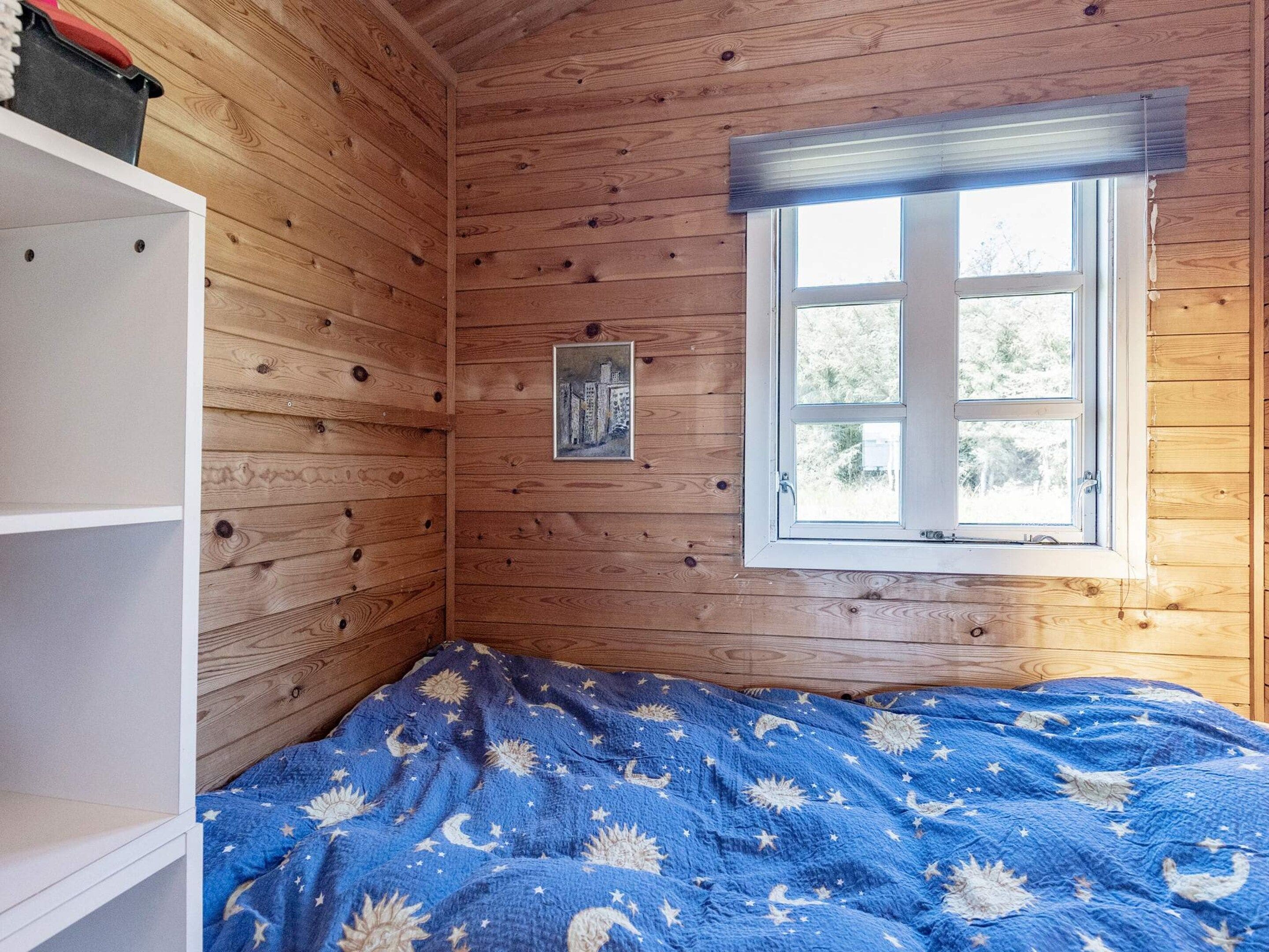 Een kamer met houten wanden, een enkel raam met zonwering, een bed met een deken met sterrenprint, en een klein schilderij aan de muur.