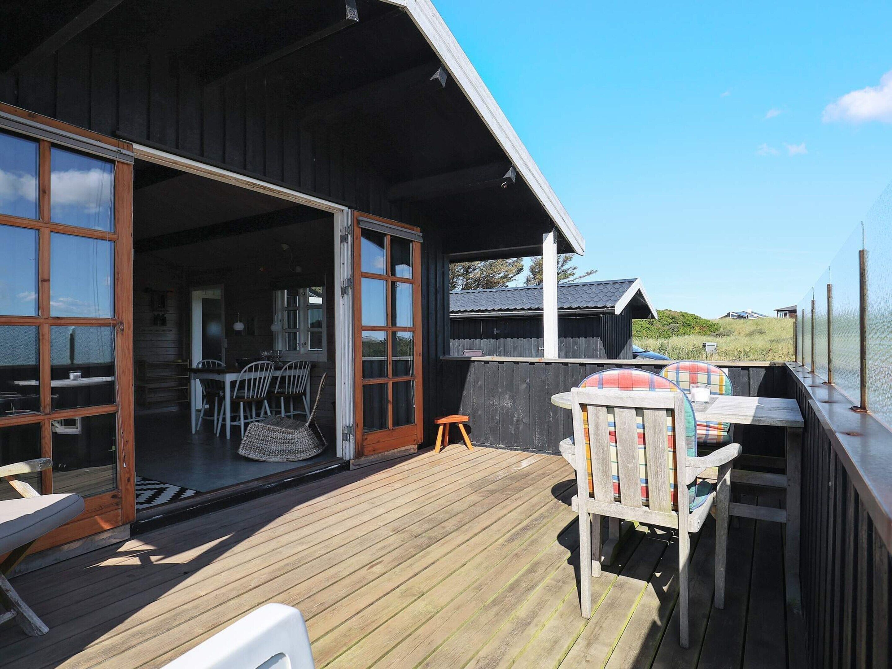 A wooden deck with outdoor furniture, a sliding glass door leading to an interior space with a dining table and chairs, and a view of the sea.