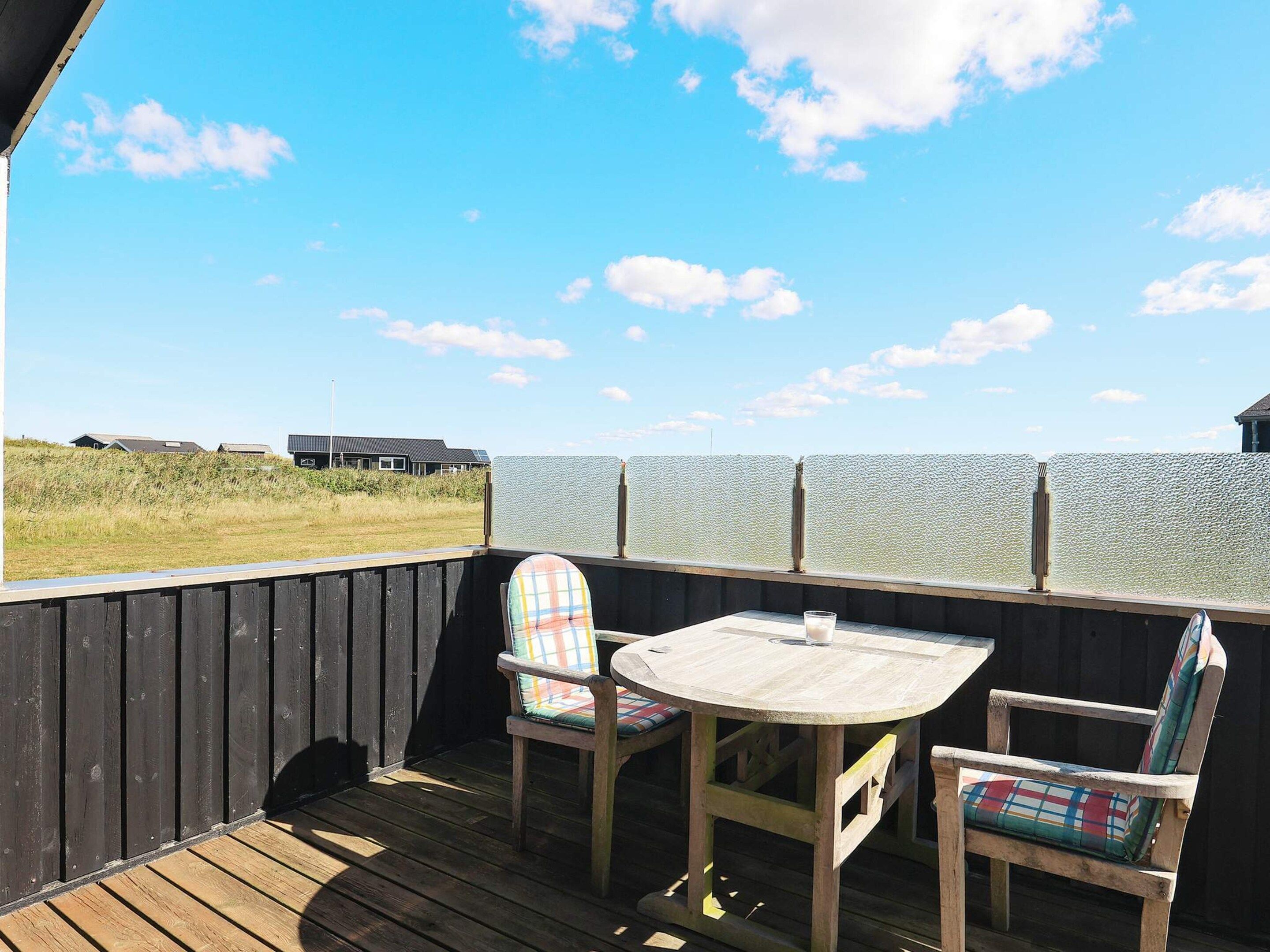 Eine Holzterrasse mit einem runden Tisch und zwei Stühlen, mit Blick auf ein Gewässer und eine Rasenfläche.
