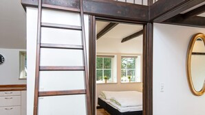 A bedroom with a bed, a wooden ladder, a window with plants, and a mirror on the wall.