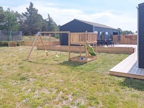 Une structure de jeux en bois comprenant un toboggan et des balançoires, une zone délimitée par une clôture, et un bâtiment doté d’un toit en tuiles.