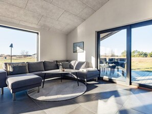 En moderne stue med en hjørnesofa, et rundt sofabord og udsigt til naturen gennem store skydedøre.