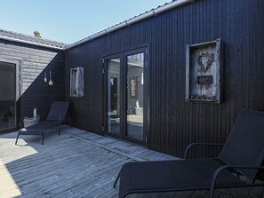 Une terrasse en bois avec deux chaises longues, une porte-fenêtre coulissante et un mur extérieur de couleur foncée agrémenté d’éléments décoratifs.