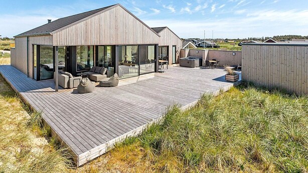 Una casa moderna con terraza de madera, amplios ventanales y mobiliario exterior.