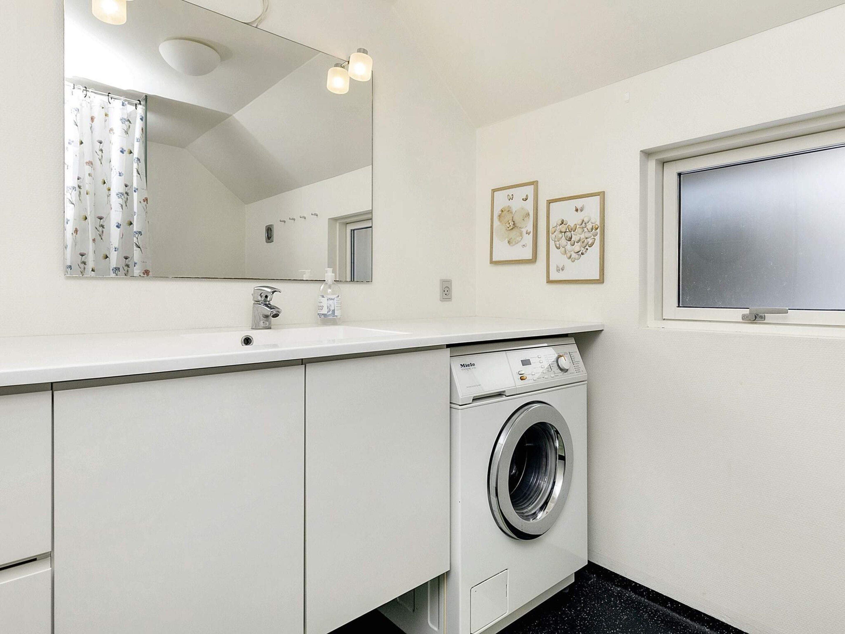 A modern bathroom with a white sink, a mirror, a washing machine, and framed artwork on the wall.