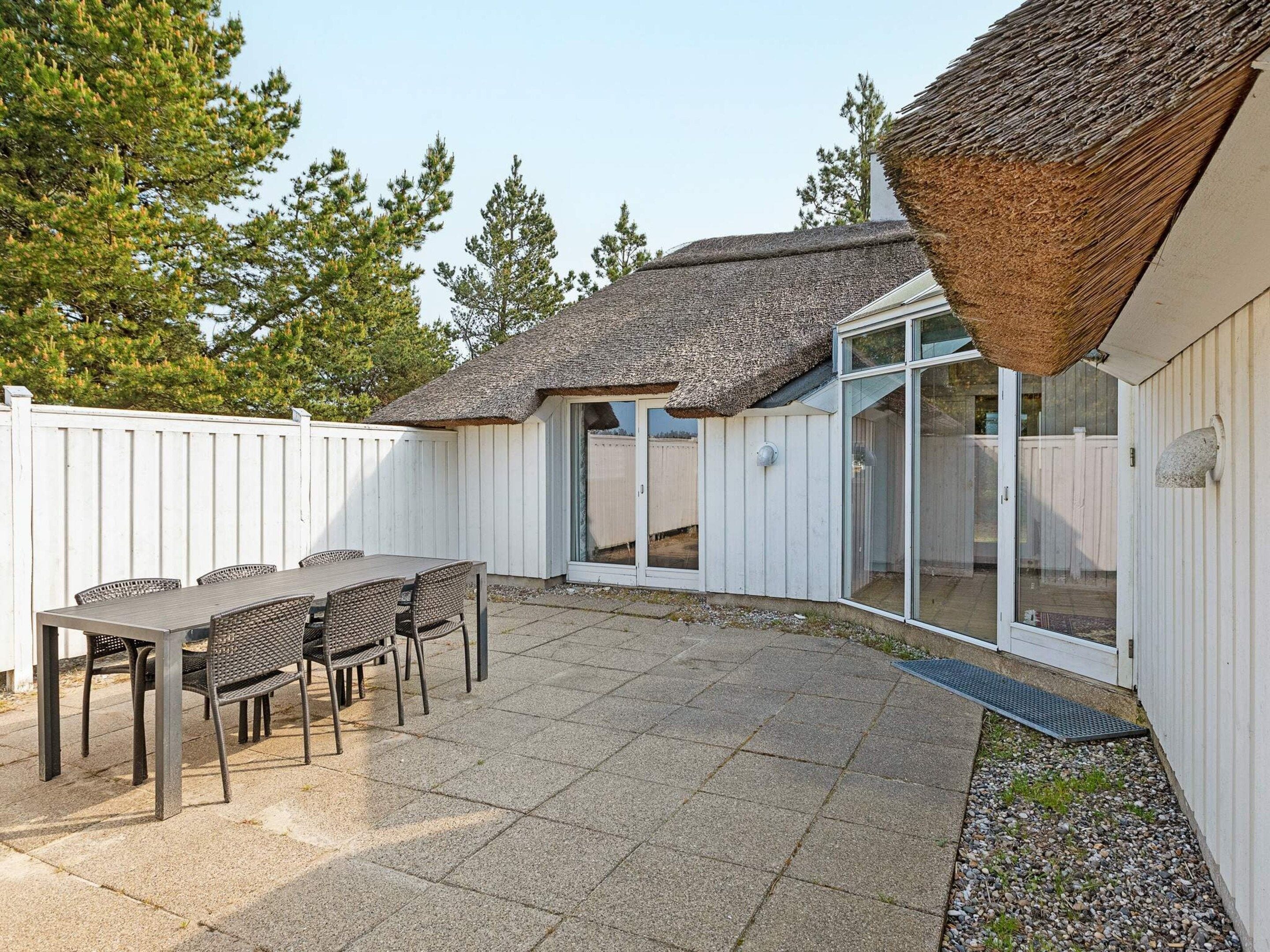 Un patio con una mesa y sillas, una estructura de techo de paja y una valla blanca.