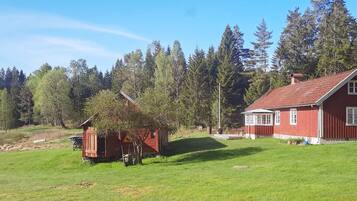 A red wooden cabin with a small porch and a larger red building with a tiled roof, surrounded by a grassy field and dense forest.