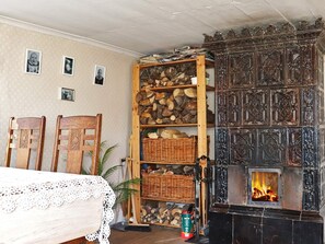 Une pièce chaleureuse avec une cheminée, des étagères en bois où sont entreposés des bûches, une nappe en dentelle et des chaises.