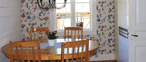 A dining area with a round wooden table, four wooden chairs with striped cushions, a floral wallpaper, a wall clock, and a window with a view of the outside.