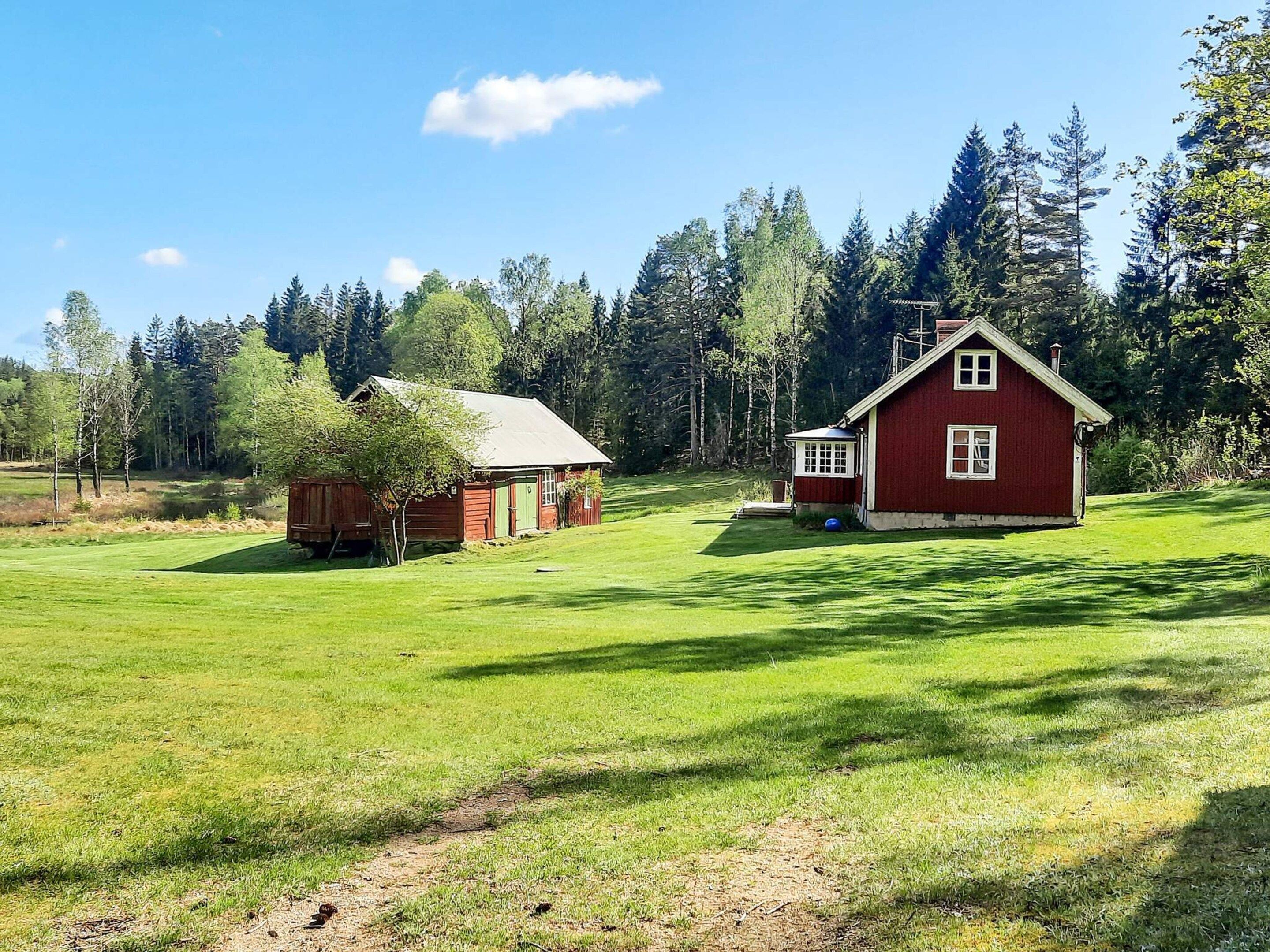 A red wooden house with a green door and a small shed in a grassy field with trees in the background.