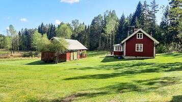A red wooden house with a green door and a small shed in a grassy field with trees in the background.