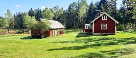 A red wooden house with a green door and a small shed in a grassy field with trees in the background.