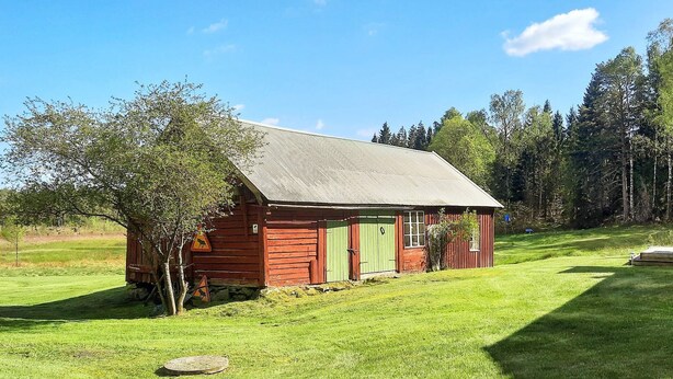 A small, single-story log cabin with a green door and a red exterior, surrounded by a grassy field and trees. - 5 Person Holiday Home in Fjaras (Fjärås)