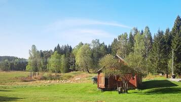 Una cabana rústica amb un teulat a pendent, envoltada per un camp verdós i un bosc dens.
