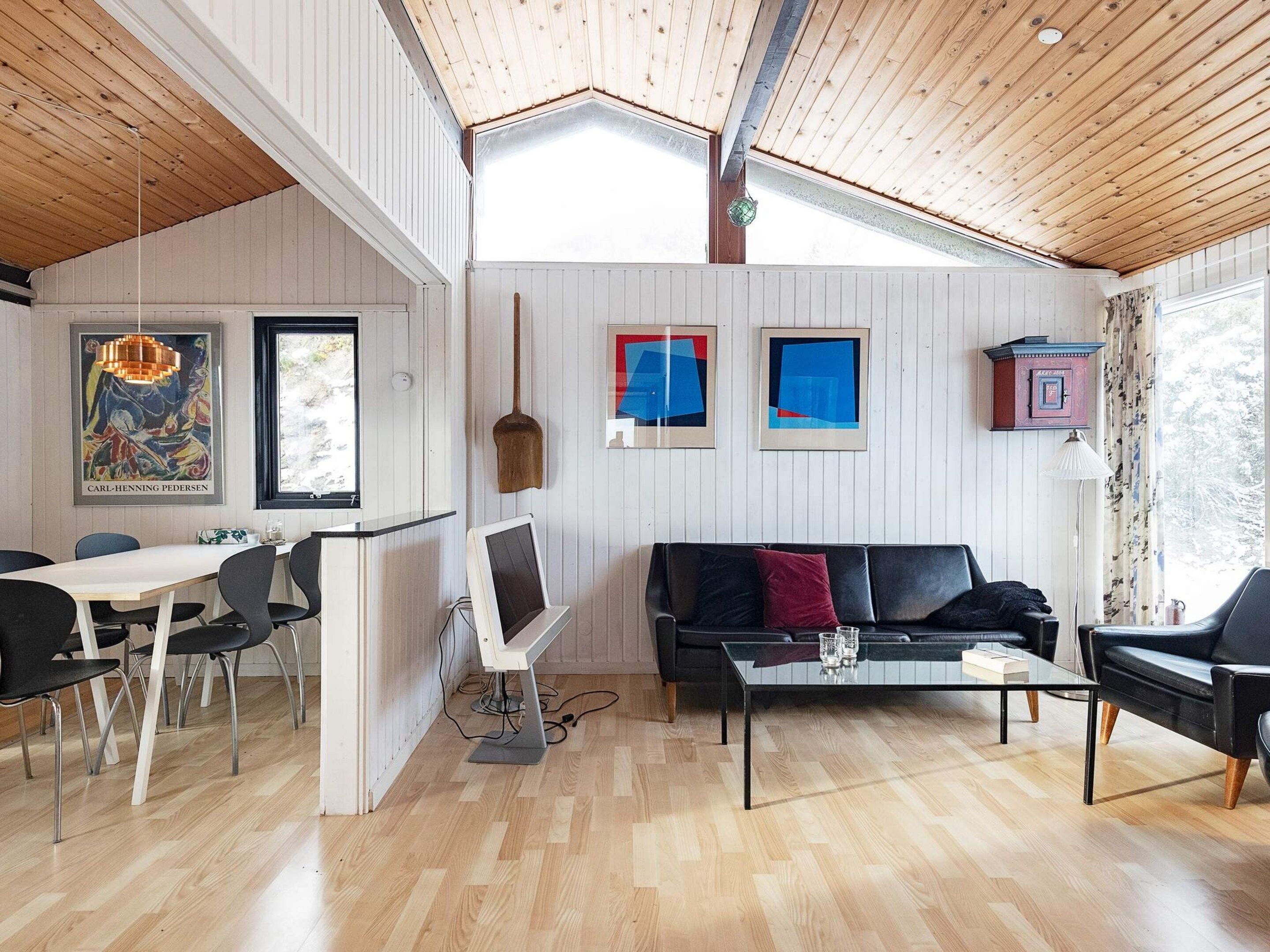 A room with a wooden ceiling, a black leather sofa, a glass coffee table, and a dining area with a white table and black chairs.