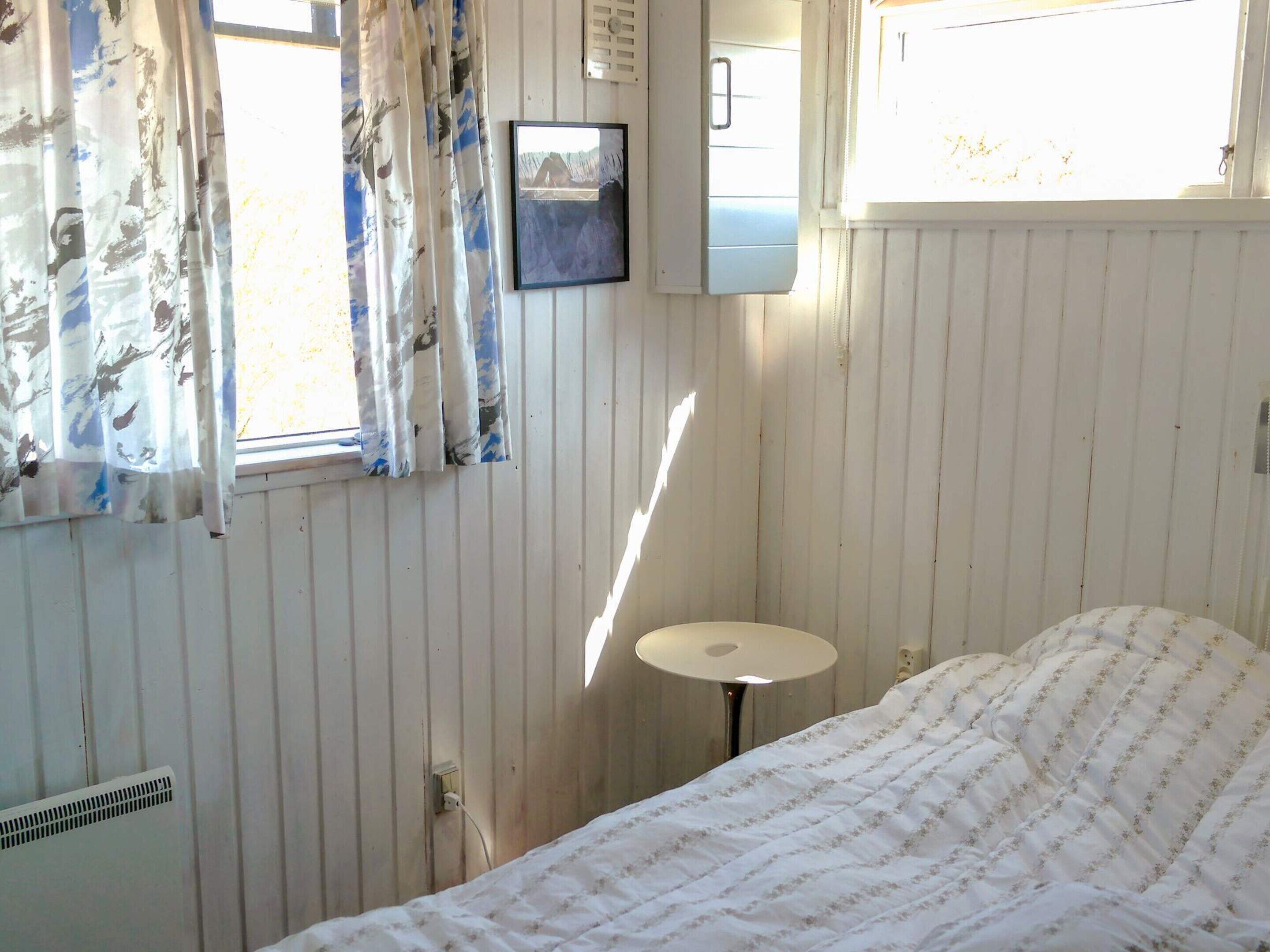 A bedroom with a bed, a small round table, a window with curtains, and a wall-mounted heater.
