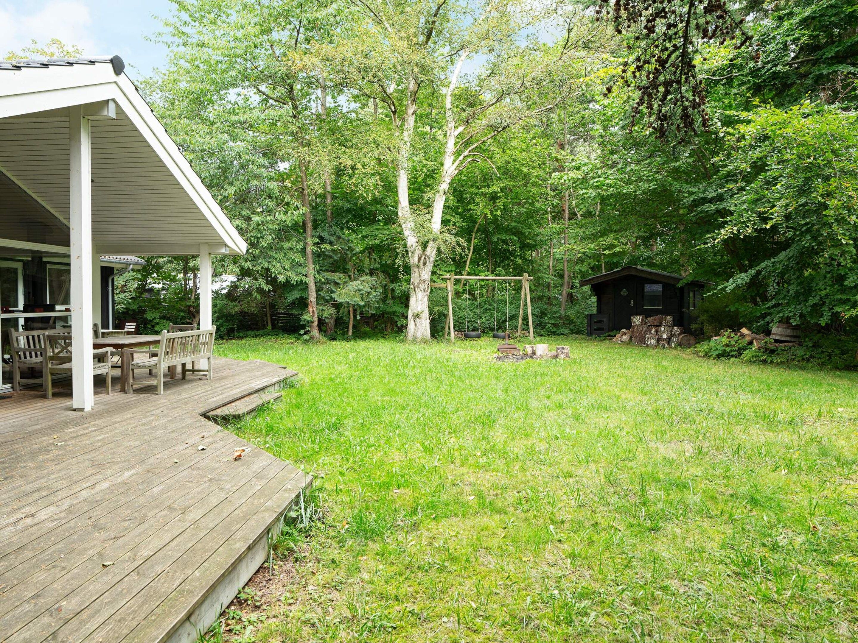 Een houten terras met tuinmeubilair, een schommelset en een kleine tuinschuur in een weelderige groene achtertuin.