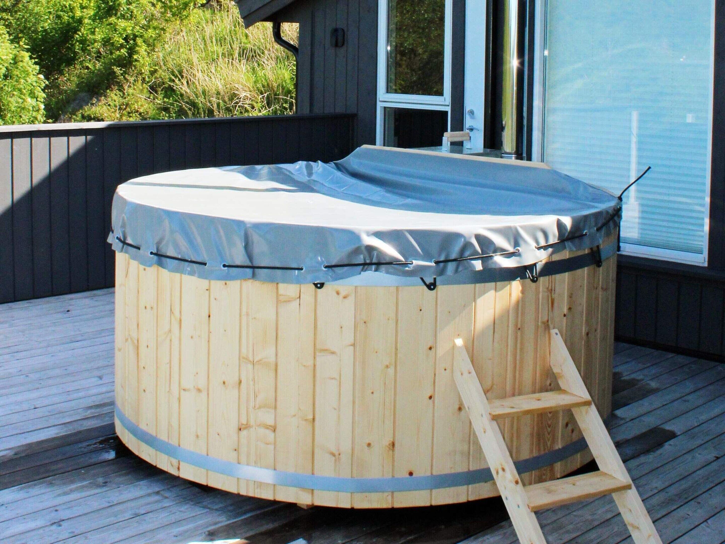 Un bain à remous en bois, avec une échelle, installé sur une terrasse en bois, entouré d’une clôture sombre et d’une maison dotée de grandes fenêtres.