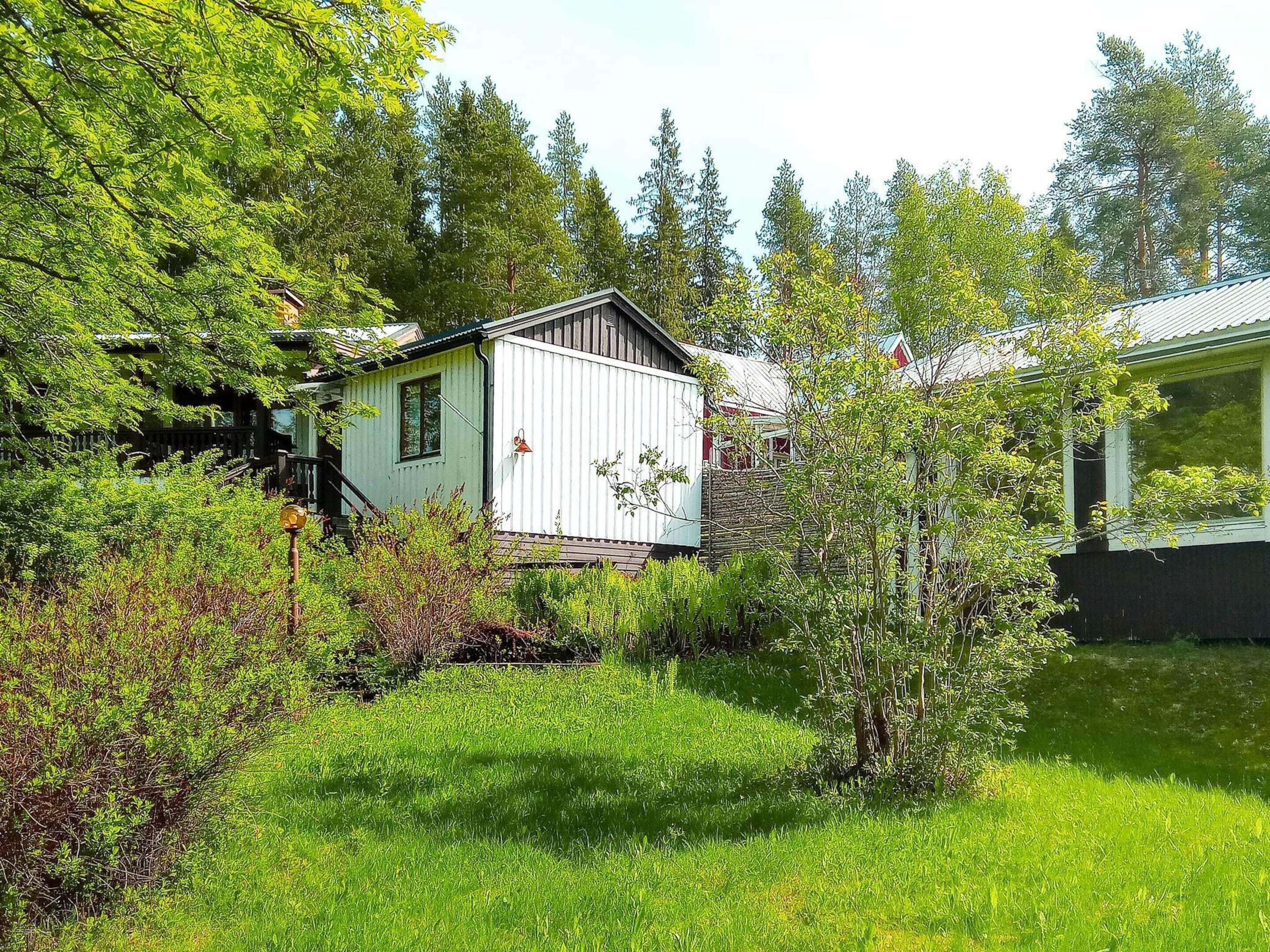 A small, single-story house with a white exterior and a dark roof, surrounded by lush greenery and trees.