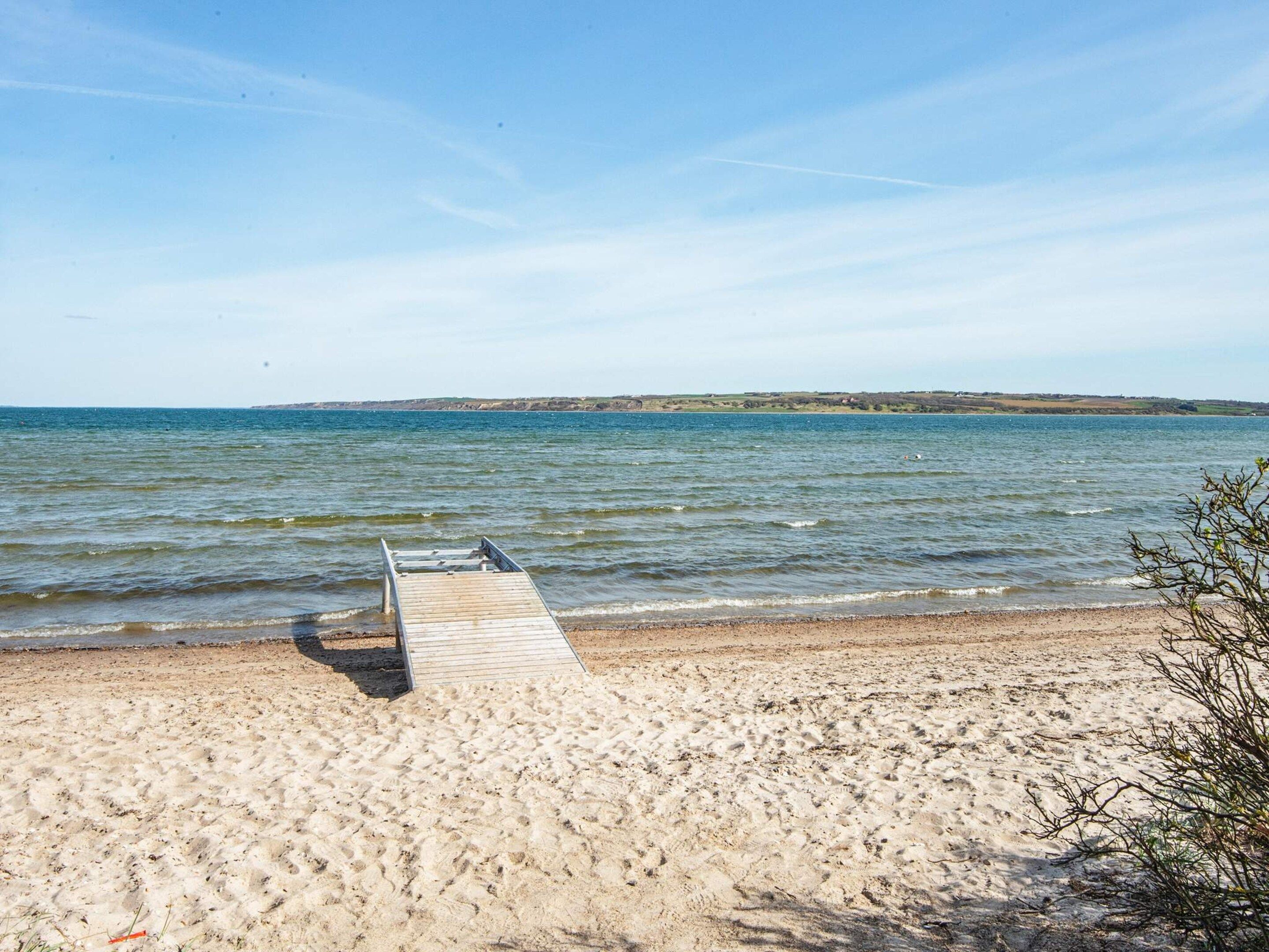 Una playa con un paseo de madera que lleva al agua, orilla arenosa y colinas a lo lejos.