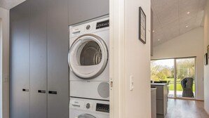 A laundry room with stacked washing machines and dryers, a door leading to a kitchen area, and a view of a garden through a glass door.
