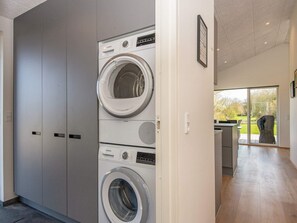 Un cuarto de lavandería con lavadoras y secadoras apiladas, una puerta que da a una zona de cocina y vistas a un jardín a través de un ventanal.