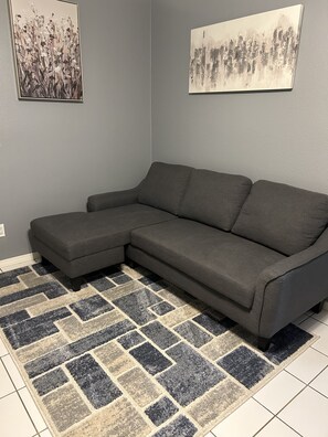A gray sectional sofa with a geometric patterned rug, two wall-mounted abstract paintings, and a tiled floor.