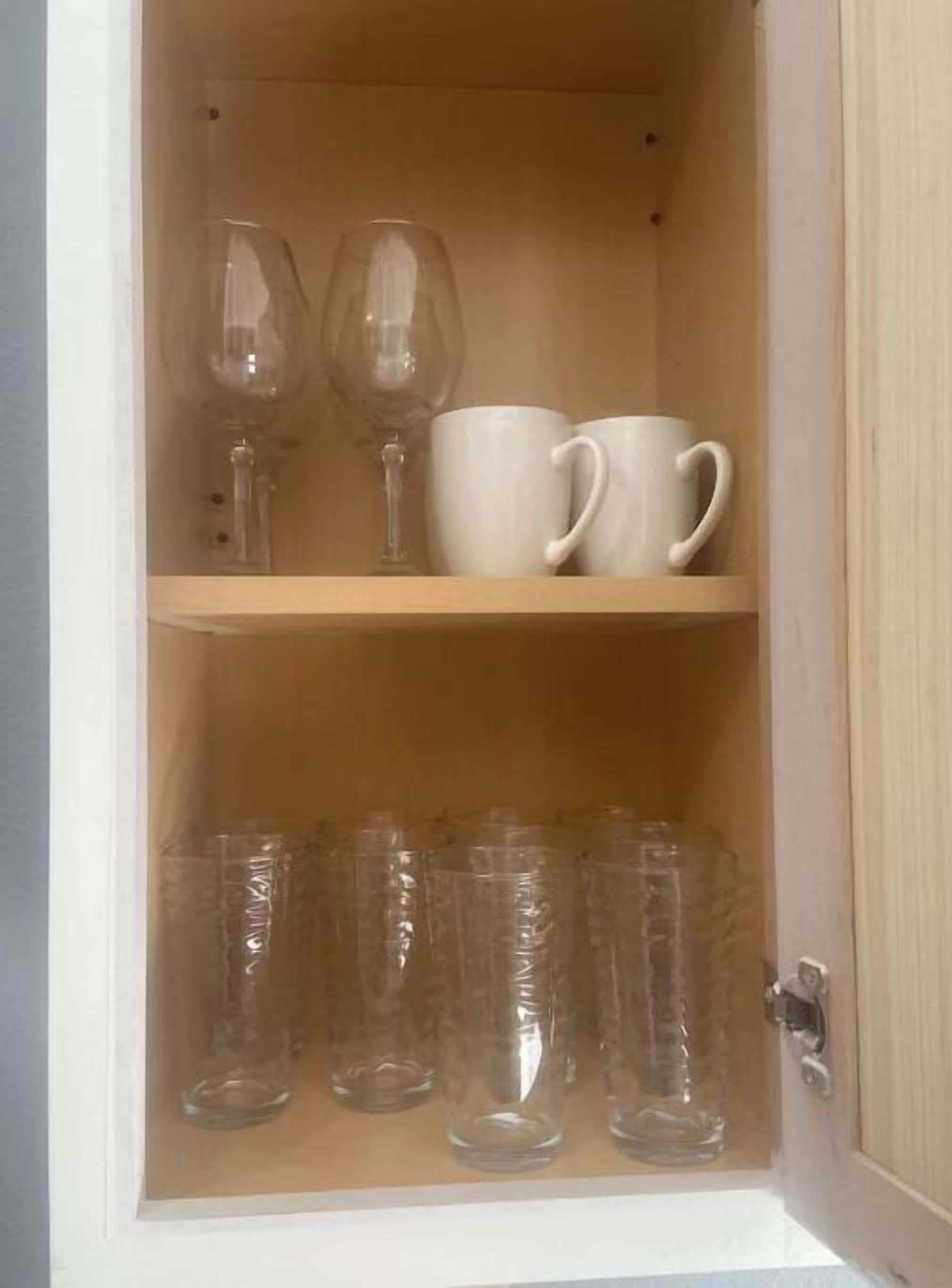 Un gabinete de madera con dos estantes; el estante superior contiene dos copas de vino y dos tazas blancas, y el estante inferior tiene varias copas de plástico transparente.